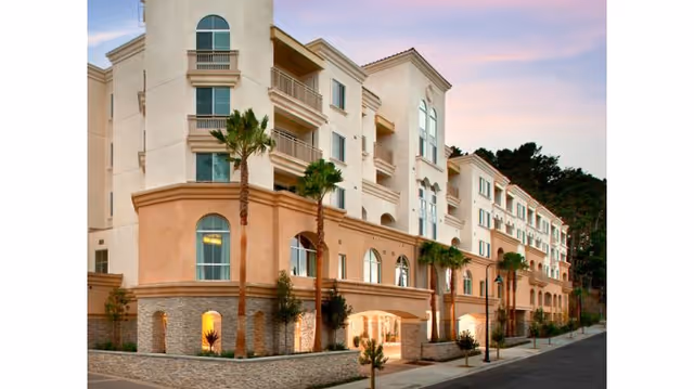 Exterior view of a multi-story senior living facility building named Peninsula Del Rey with palm trees lining the sidewalk and a clear sky at dusk.