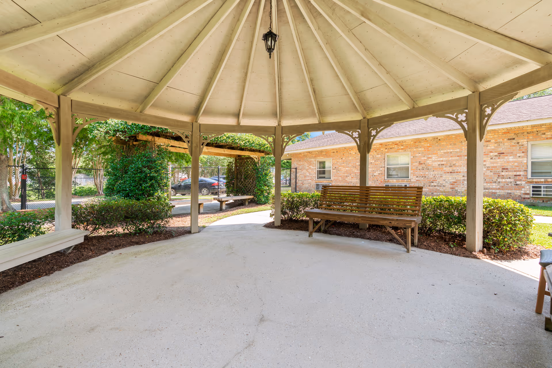 View from inside a covered outdoor gazebo with a wooden bench and white ceiling. Surrounding the gazebo are bushes, trees, and a brick building with windows. A black car is parked outside near a fence.