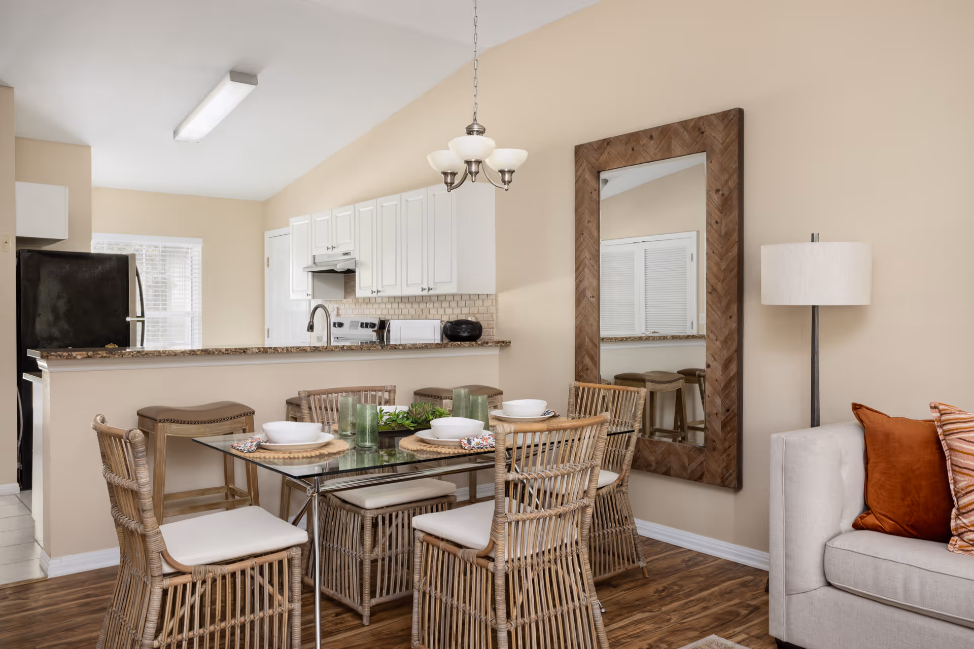 A cozy dining area with a glass-top table surrounded by six wicker chairs with white cushions. The table is set with white bowls, plates, green glasses, and woven placemats. Behind the dining area is a kitchen with white cabinets, a black refrigerator, and a granite countertop with two bar stools. A large wooden-framed mirror hangs on the beige wall next to a floor lamp and a light gray sofa with orange and striped pillows.
