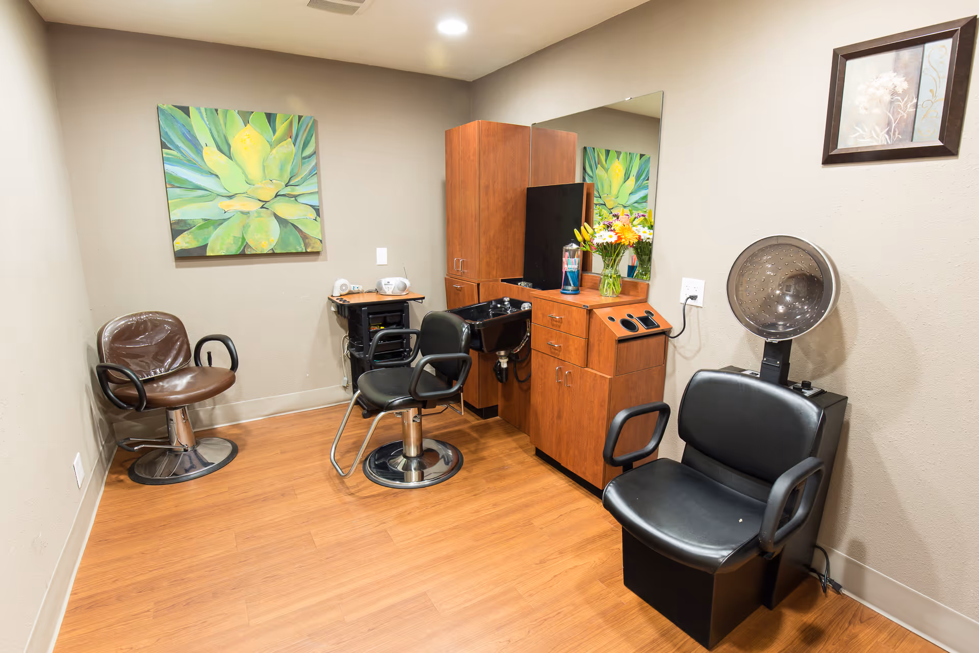 Interior view of a small hair salon room with three salon chairs, a hair washing station, wooden cabinets, a large mirror, and decorative wall art. The room has wood flooring and neutral-colored walls.