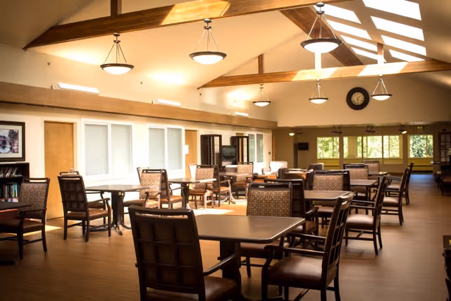 A spacious dining room with multiple tables and chairs arranged neatly. The room features a high ceiling with wooden beams and hanging light fixtures. Large windows allow natural light to fill the space, and a clock is mounted on the far wall above the windows.