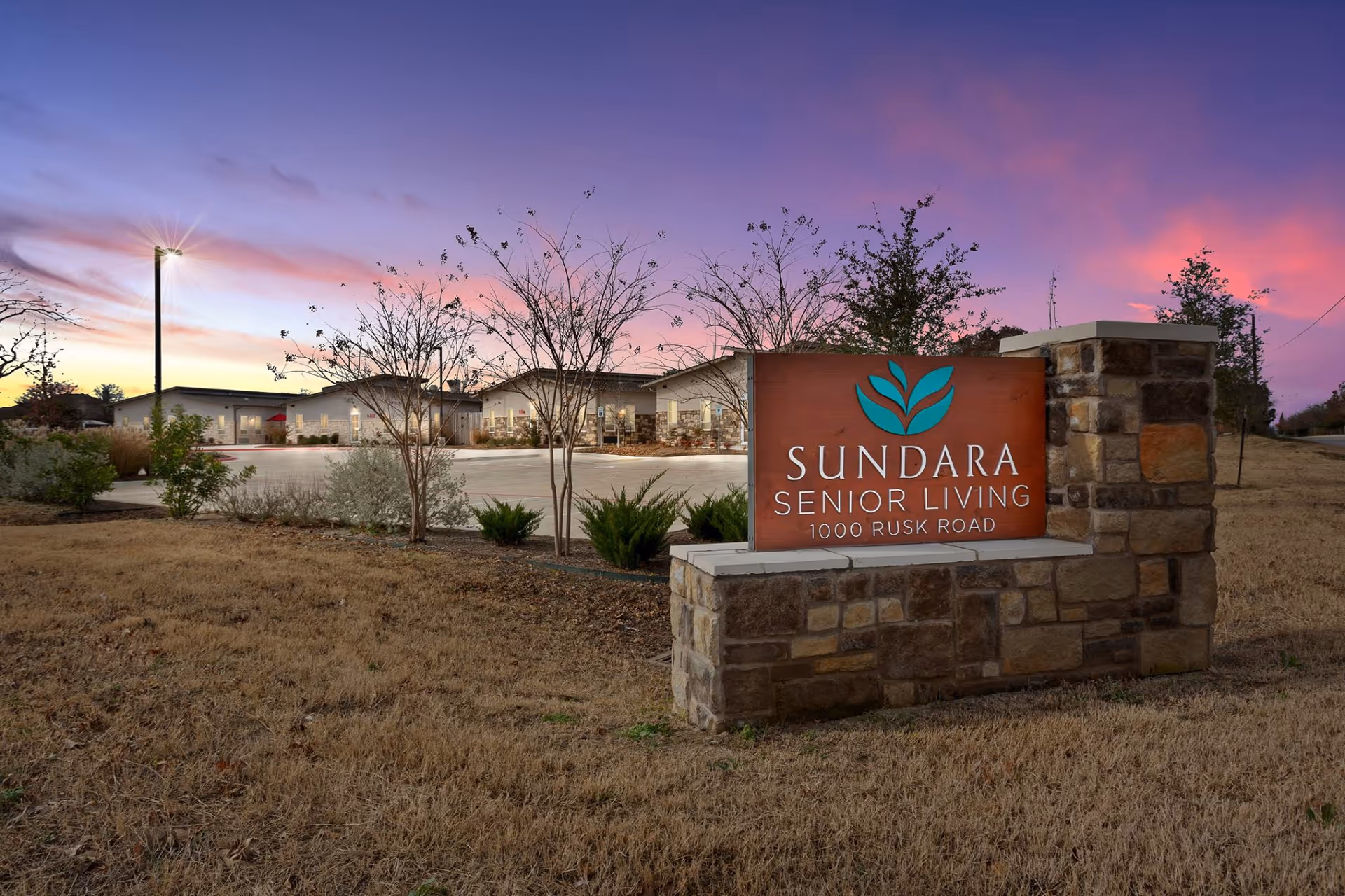 Stone sign reading 'Sundara Senior Living 1000 Rusk Road' stands in front of the facility at dusk with the building and parking area behind it.