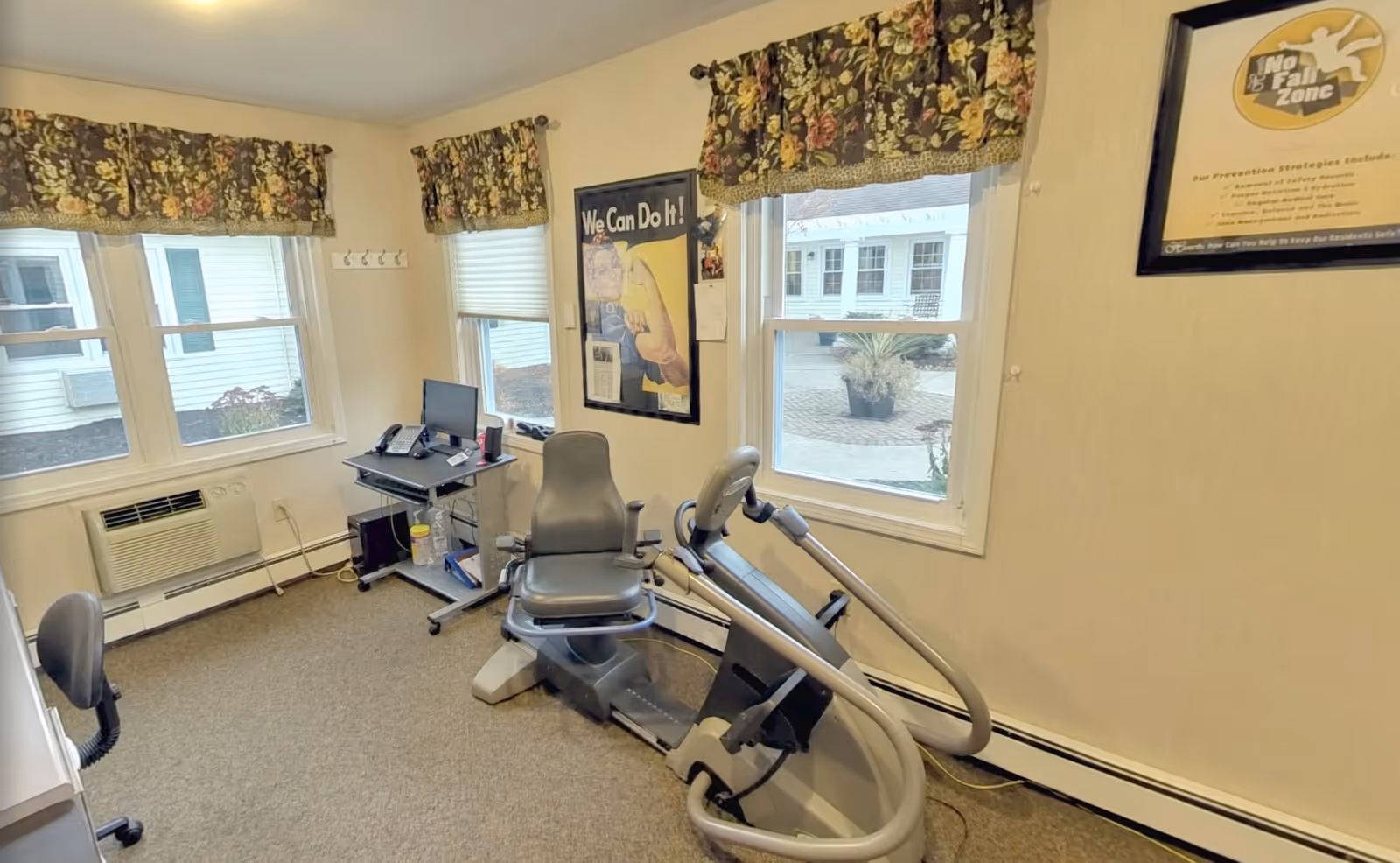 A small exercise room with a recumbent exercise bike, two office chairs, a small desk with a computer and phone, and three windows with floral valances. The walls are light-colored and decorated with framed posters, including a 'We Can Do It!' poster and a 'No Fall Zone' safety poster.