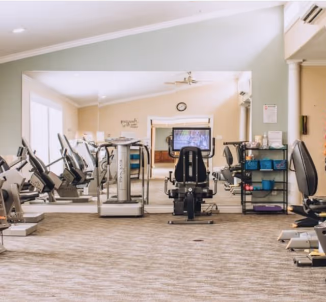 A fitness room with several exercise machines including stationary bikes and a recumbent bike facing a large mirror. The room has beige walls, carpeted floor, a clock on the wall, and a small shelving unit with exercise accessories.