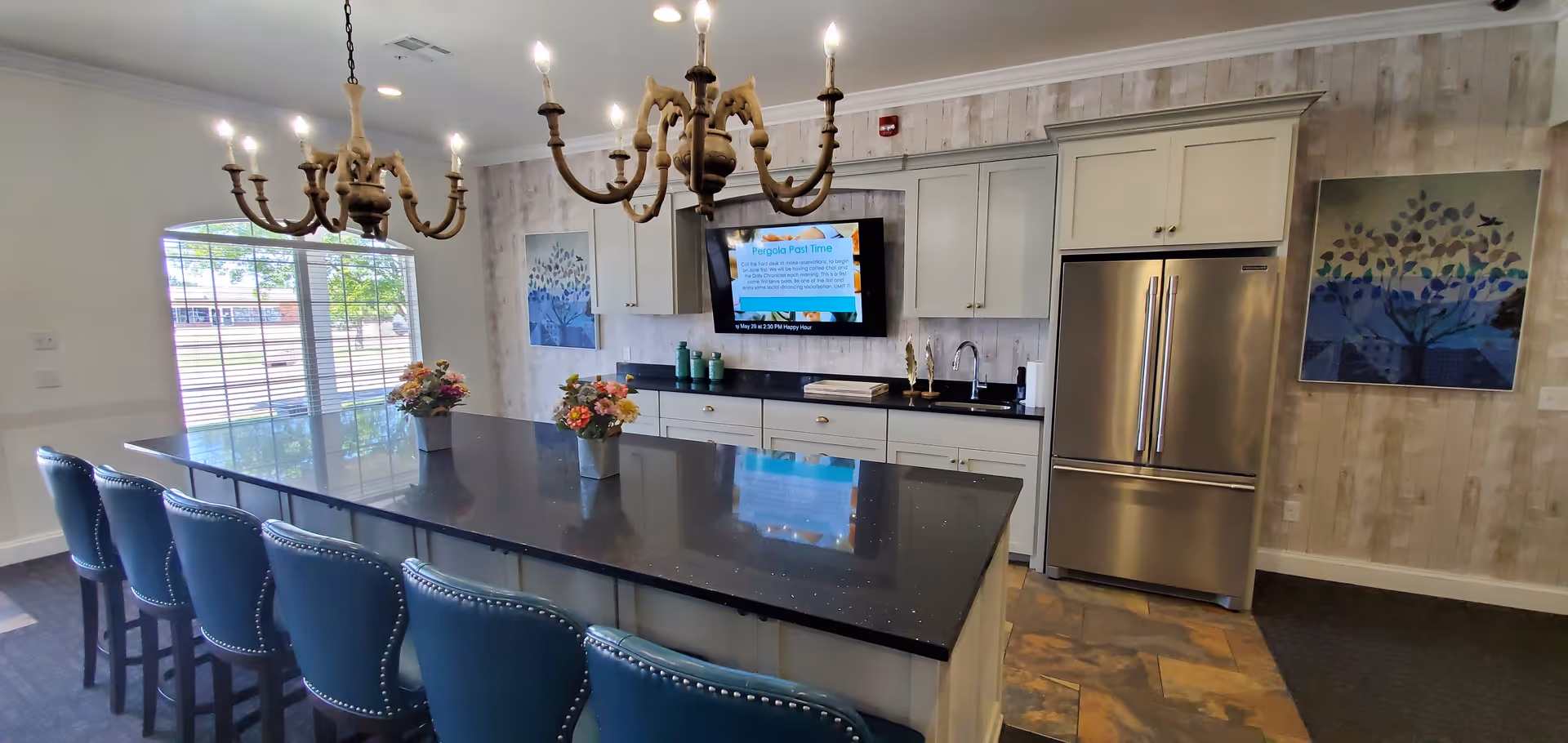 Modern kitchen area in Hefner Mansions Senior Independent Living featuring a large black granite island with six blue cushioned bar stools, two ornate chandeliers hanging above, white cabinetry, stainless steel refrigerator, a mounted TV, and decorative artwork on the walls.