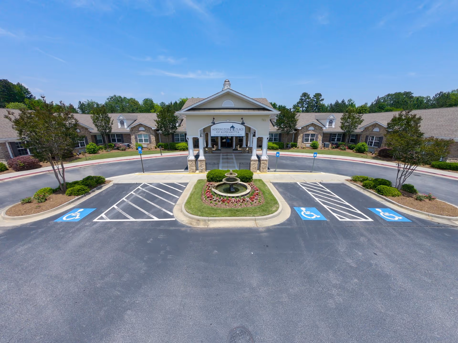 Front exterior view of Addington Place Of Alpharetta, showing a large circular driveway with handicap parking spaces, a central landscaped area with a fountain, and a covered entrance to the building. The building is surrounded by trees and shrubbery under a clear blue sky.