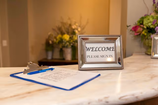 A reception desk with a clipboard holding a sign-in sheet and a pen. A framed sign on the desk reads 'WELCOME PLEASE SIGN IN'. In the background, there are blurred flower arrangements on a table against a beige wall.