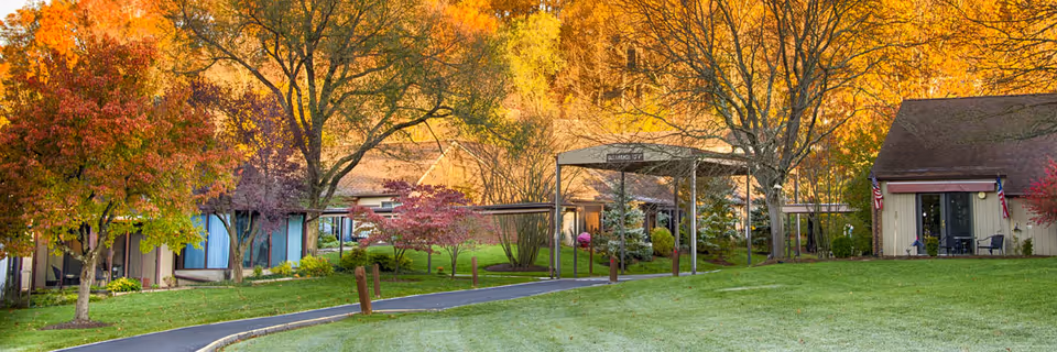 A scenic outdoor view of a senior living facility named Broadmead, featuring a paved walkway, green lawn, and trees with autumn-colored leaves. Several single-story buildings with sloped roofs and American flags are visible, surrounded by well-maintained landscaping.