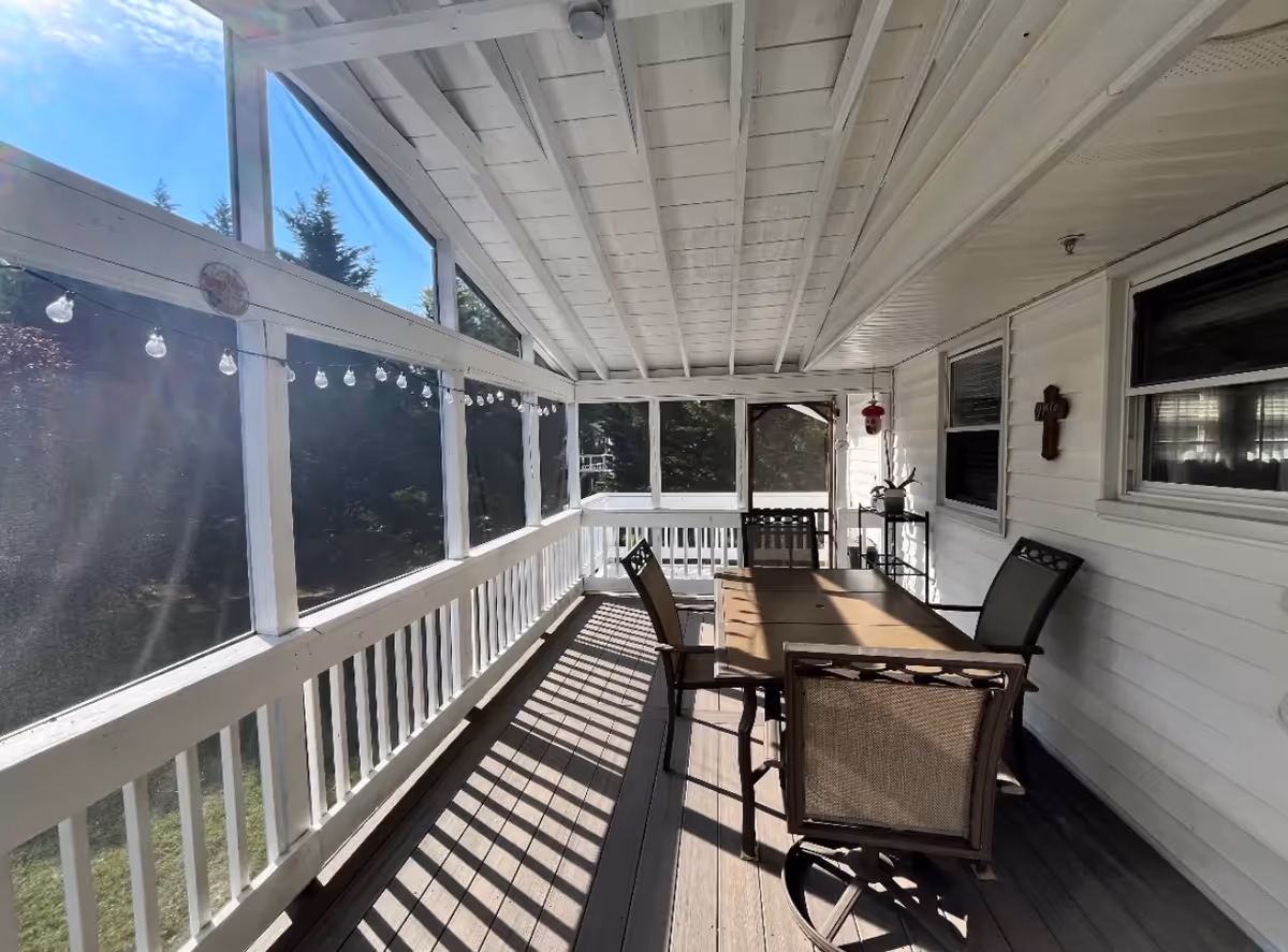 A covered porch area with a wooden table and four chairs. The porch has white railings and a white ceiling with exposed beams. String lights are hung along the windows, and there is a small shelf with a plant and a decorative cross on the wall. Trees are visible outside the screened windows.