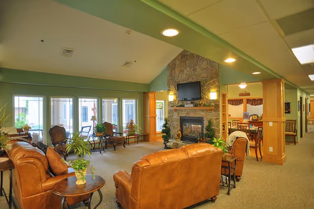 A cozy living room area in a senior living facility with brown leather sofas, a stone fireplace with a mounted TV above it, several potted plants, and large windows letting in natural light. Adjacent to the living room is a dining area with tables and chairs.
