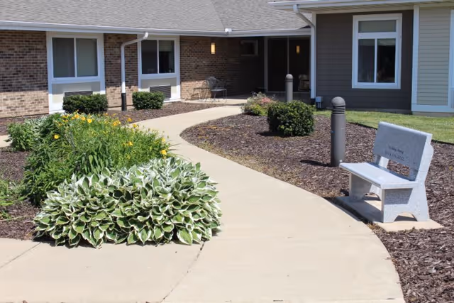A curved concrete walkway leading to the entrance of a brick and siding building, surrounded by landscaped garden beds with green shrubs and yellow flowers. A gray memorial bench is placed on the right side of the walkway.
