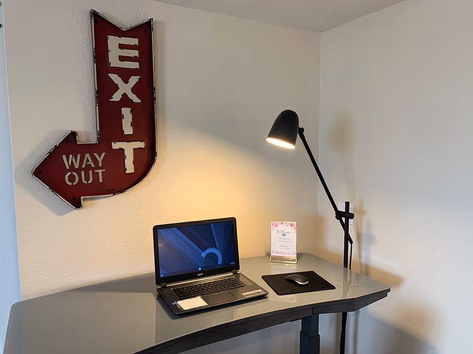 A modern workspace with a gray desk holding a laptop, a mouse on a black mousepad, a small welcome sign, and a black adjustable desk lamp. On the wall above the desk is a red vintage-style metal sign shaped like an arrow pointing left with the words 'EXIT WAY OUT'.