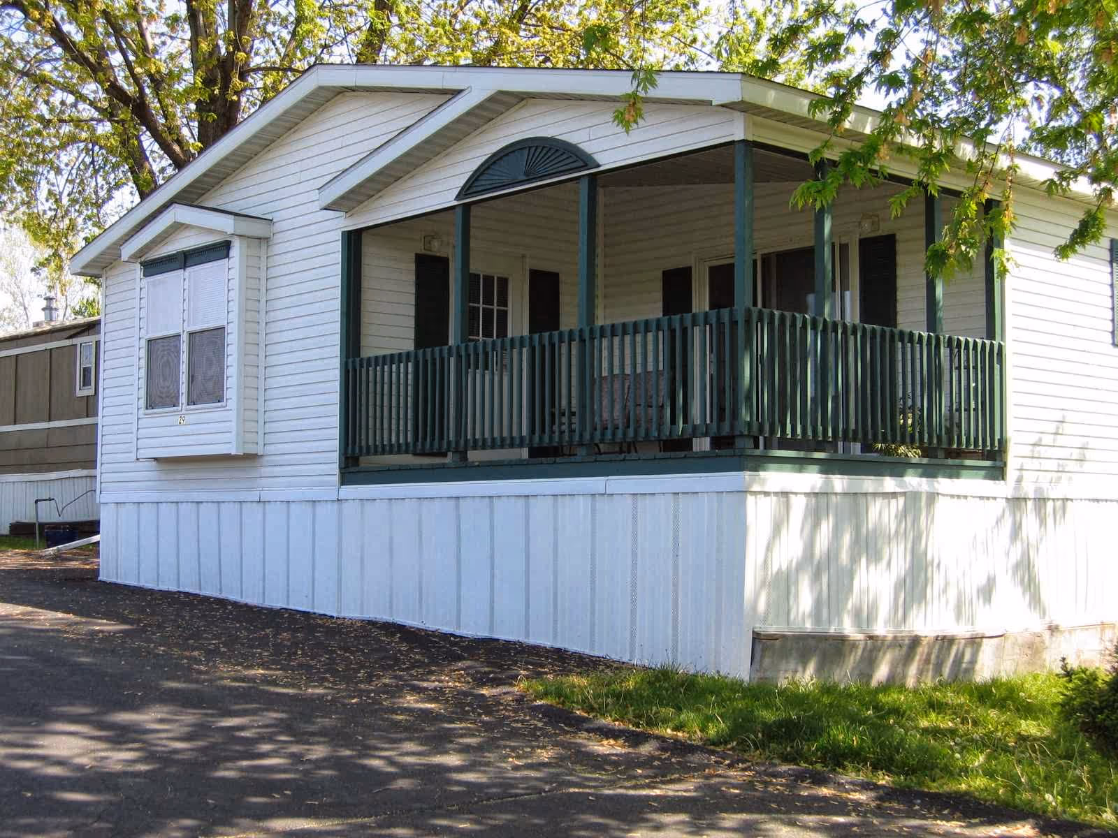 Front exterior of a white mobile home with a covered green-railed porch and nearby trees.