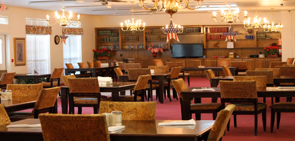 A spacious dining room with multiple tables and upholstered chairs arranged neatly. The room has a red carpet and is decorated with patriotic star ornaments hanging from the ceiling. There are chandeliers providing warm lighting, large windows with floral valances, and a wooden bookshelf with books and a television in the background.