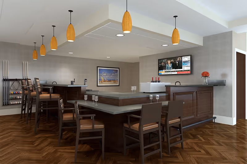 A modern indoor lounge area with a large L-shaped bar counter surrounded by wooden chairs with cushioned seats. The bar has a gray countertop and dark wood paneling. Above the bar, there are six pendant lights with wooden shades. On the wall behind the bar, a flat-screen TV is mounted showing a news broadcast. A framed picture hangs on the adjacent wall, and a rack with billiard cues and balls is visible on the left side. The floor has a herringbone wood pattern.