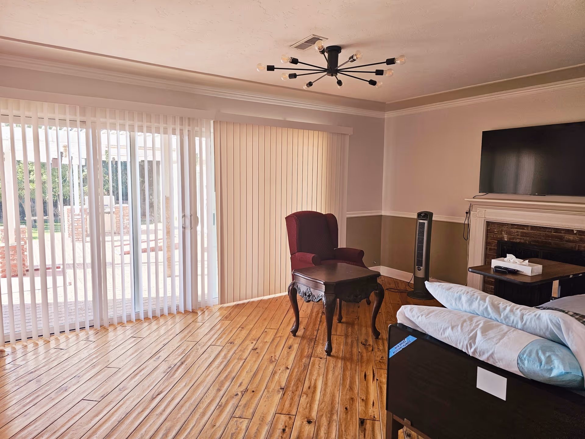 A bright room with wooden flooring featuring a large sliding glass door with vertical blinds leading to an outdoor patio. Inside, there is a maroon armchair next to a dark wooden side table, a flat-screen TV mounted above a white fireplace, a standing fan, and a hospital-style bed with pillows and a remote control on a small table beside it.