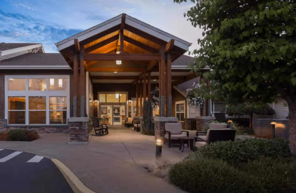 Entrance of a senior living facility with a covered porch supported by wooden beams. There are outdoor chairs and tables arranged on the right side, surrounded by greenery and trees. The building has large windows and stone accents on the lower walls. The sky is partly cloudy during dusk.