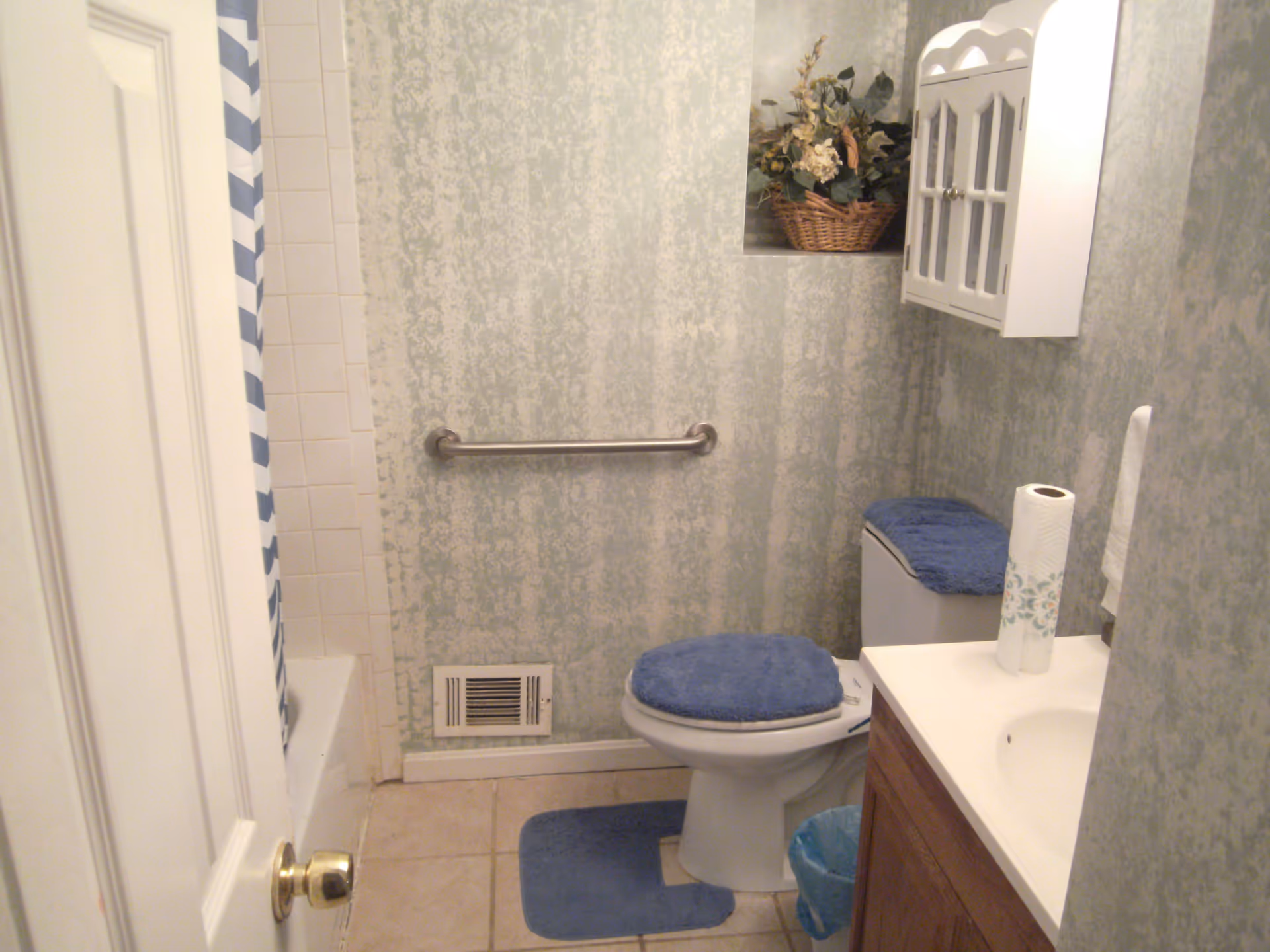 Small bathroom with a white toilet covered with a blue seat cover and a matching blue rug in front. There is a white sink with a wooden cabinet below and a roll of paper towels on the counter. A white wall cabinet with glass doors is mounted on the wall above the toilet. A basket with artificial flowers is placed in a recessed wall niche. The walls have a textured light green and white pattern. A metal grab bar is installed on the wall next to the toilet. A shower with a white tiled wall and a blue and white striped shower curtain is partially visible.