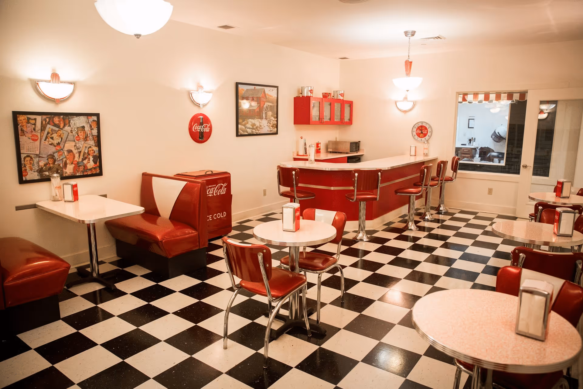 A retro-style diner interior with black and white checkered floor tiles, red and white booths and chairs, a red counter with bar stools, vintage Coca-Cola decor, and framed pictures on the walls.