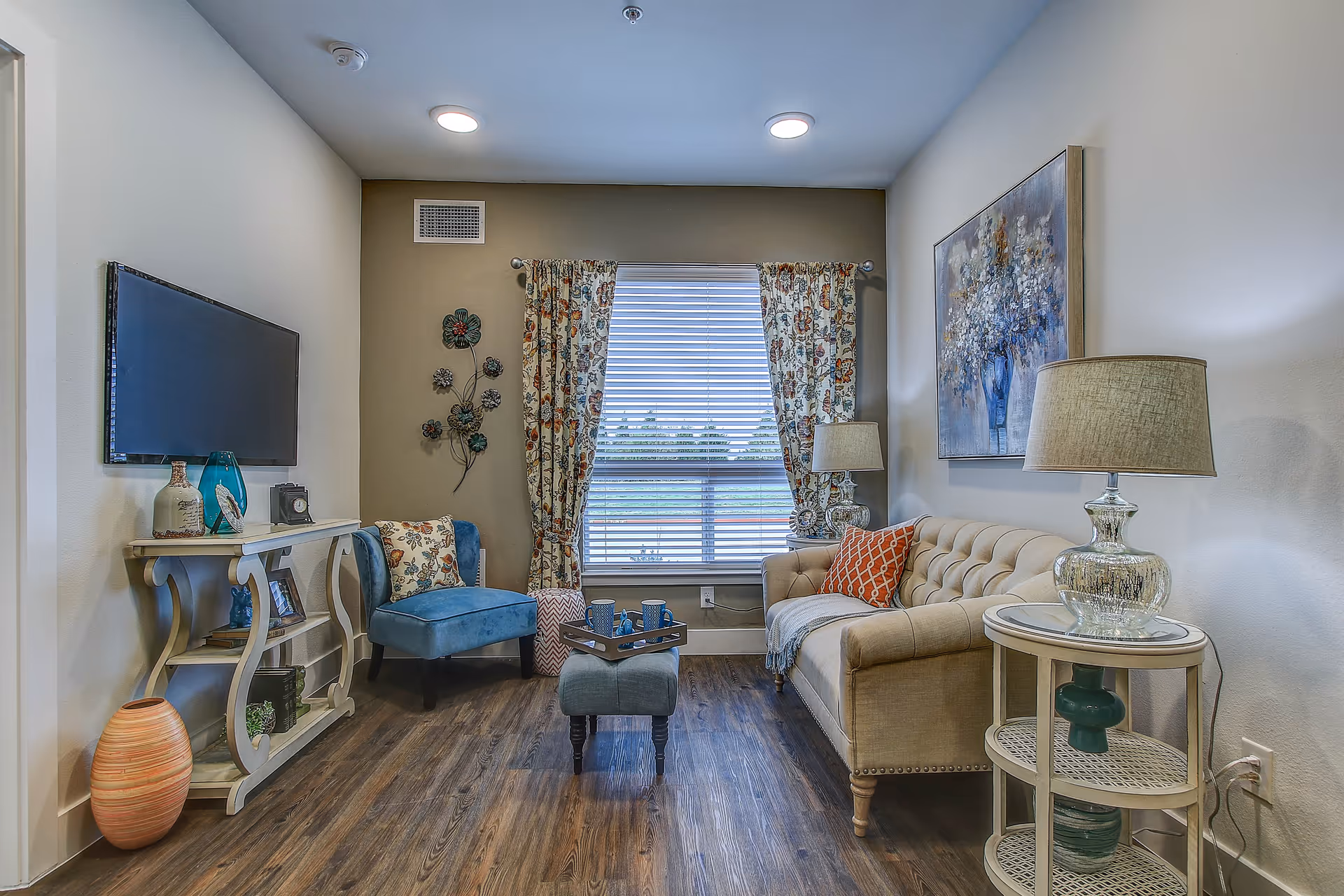 A cozy living room with a beige tufted sofa adorned with orange and beige pillows, a blue upholstered chair with a floral pillow, a small ottoman with a tray holding cups, a wall-mounted flat-screen TV, decorative wall art, floral curtains on a window, and two lamps on side tables. The room has wooden flooring and neutral-colored walls.