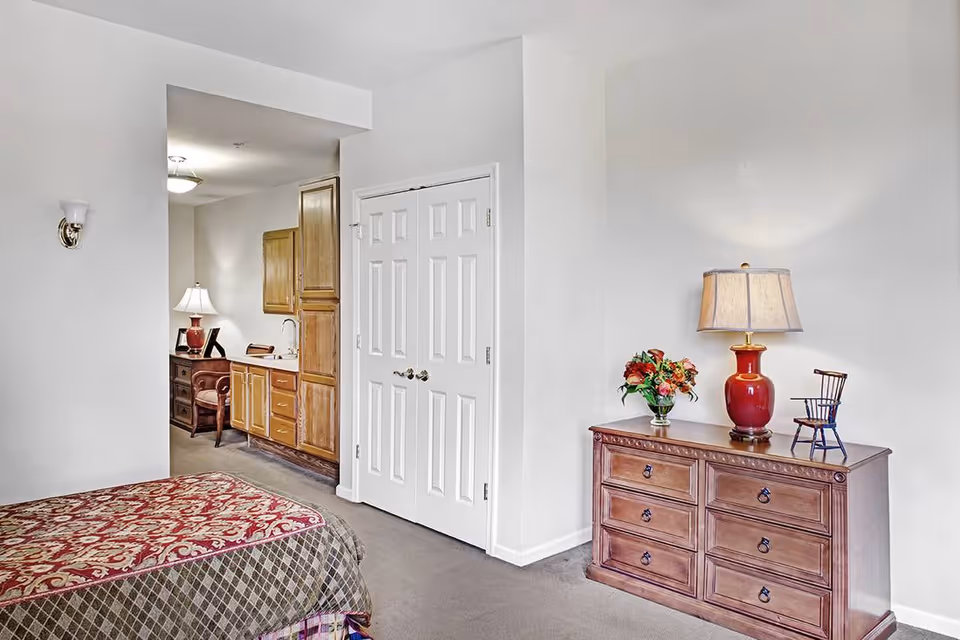 A bedroom in a senior living facility featuring a bed with a patterned red and beige bedspread, a wooden dresser with a red lamp, a small decorative chair, and a vase of flowers. In the background, there is a small kitchenette area with wooden cabinets, a sink, and a desk with a chair and lamp.