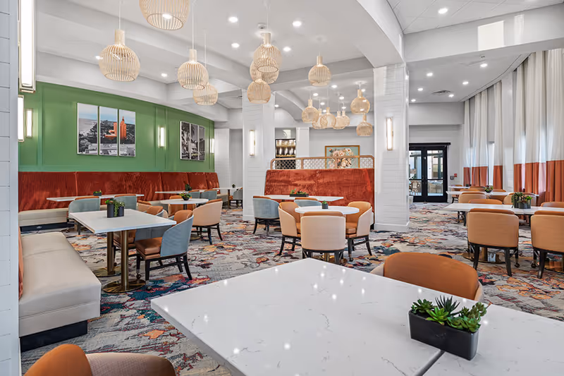 A spacious and modern dining room with multiple tables and chairs arranged neatly. The room features a colorful patterned carpet, green accent wall with framed pictures, white walls with vertical lighting fixtures, and several hanging wooden pendant lights. There are large windows with white and orange curtains allowing natural light to enter.