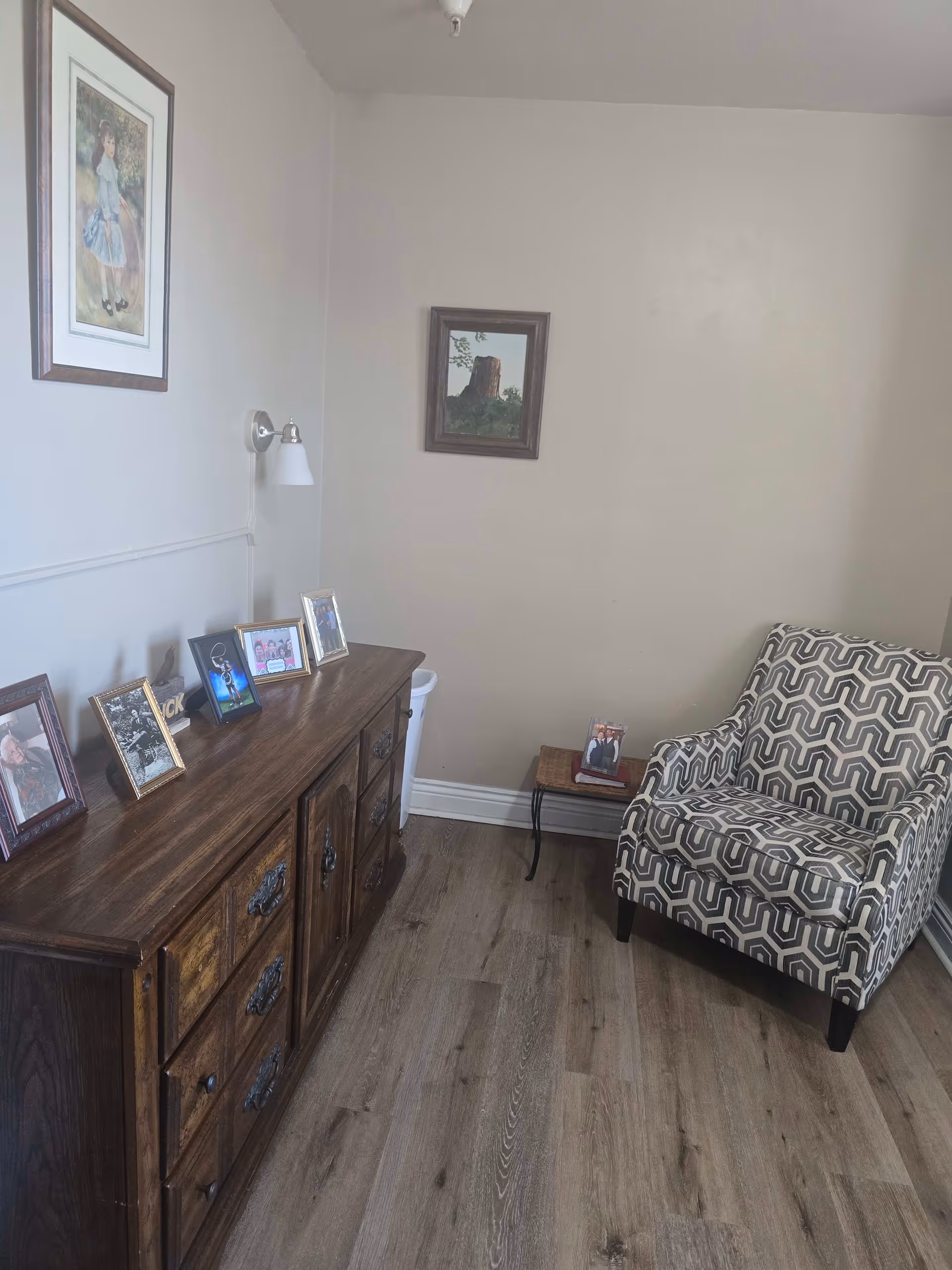 A cozy corner of a room featuring a patterned armchair, a small side table with a framed photo, and a wooden dresser with multiple framed photos on top. The walls are light-colored with two framed pictures hanging, and the floor is wood.
