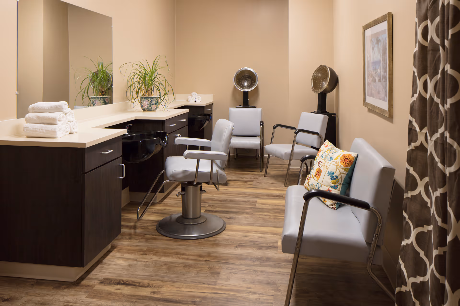 Interior view of a salon area in a senior living facility with a styling chair in front of a counter with a sink, two hair dryers on stands, a bench with a colorful pillow, and a few chairs. The room has wood flooring, beige walls, a large mirror, and a patterned curtain.