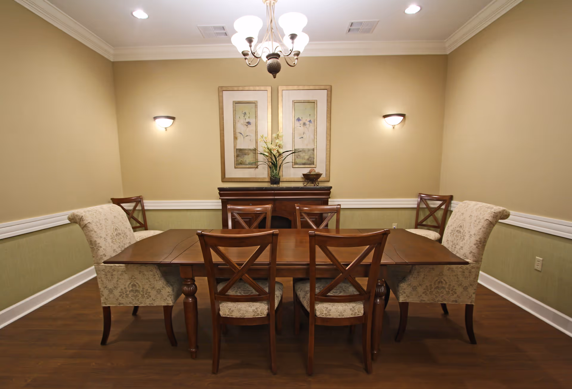 A dining room with a wooden table surrounded by six chairs, two of which have upholstered patterned fabric. The room has beige walls with white trim, two framed floral artworks on the wall, a wooden sideboard beneath the artwork, and a chandelier hanging from the ceiling. Two wall sconces provide additional lighting.