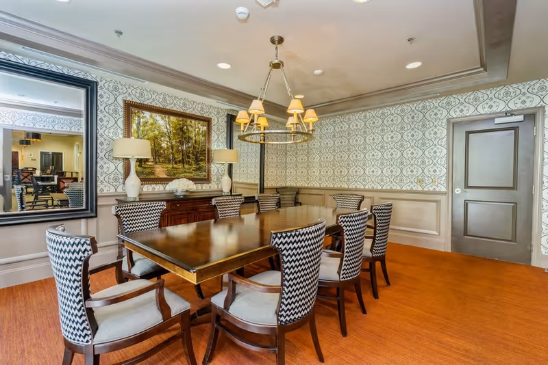 A formal dining room with a long wooden table surrounded by eight upholstered chairs with a black and white zigzag pattern. The room features patterned wallpaper, a large mirror, a framed landscape painting, two table lamps on a wooden sideboard, and a chandelier with multiple lampshades hanging above the table. The floor is covered with a warm-toned carpet, and there is a closed gray door on the right side.