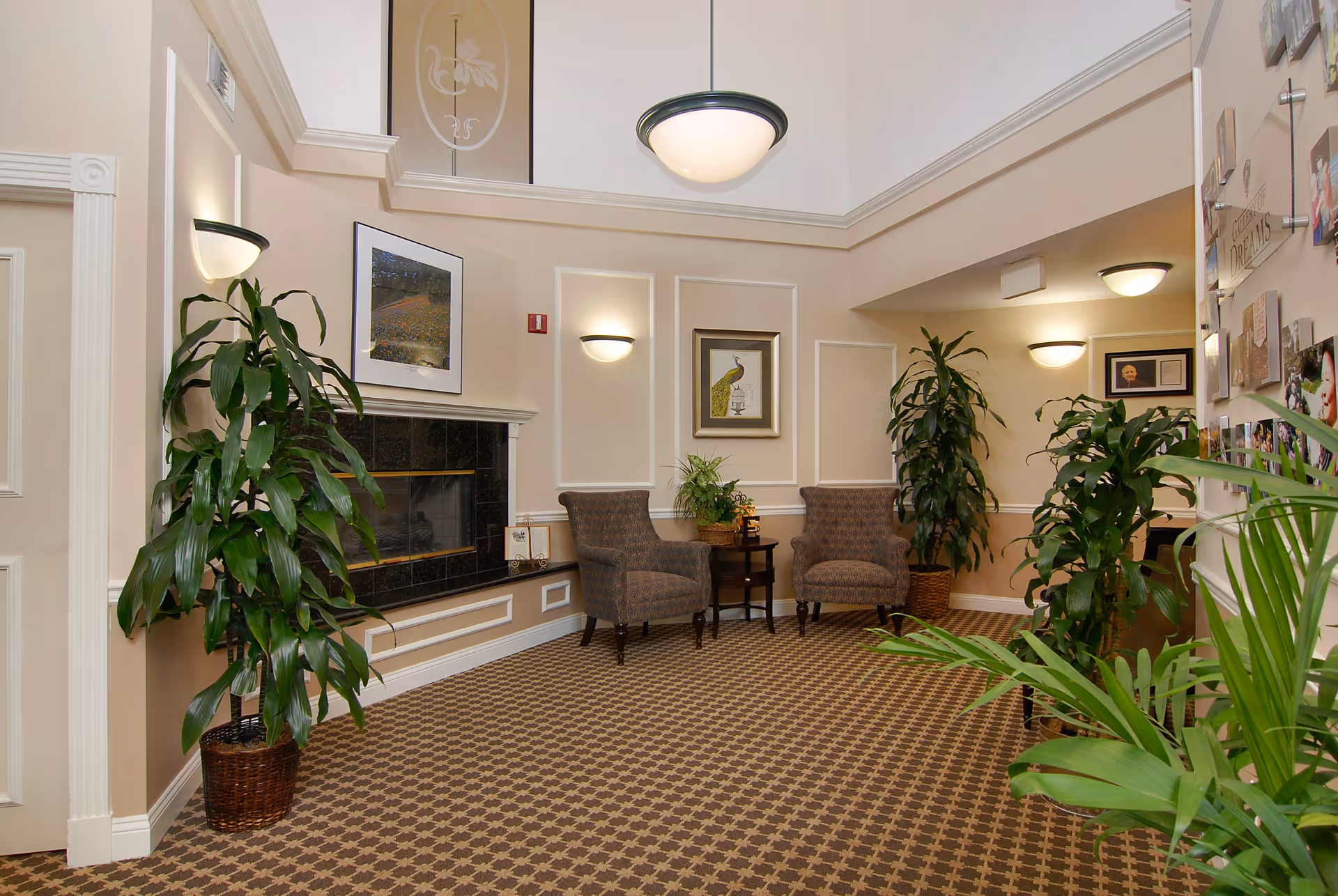 A cozy interior seating area with two upholstered armchairs and a small table between them, surrounded by several large potted plants. The room features a fireplace with a framed picture above it, beige walls with white trim, patterned carpet, and soft wall and ceiling lights providing warm illumination.