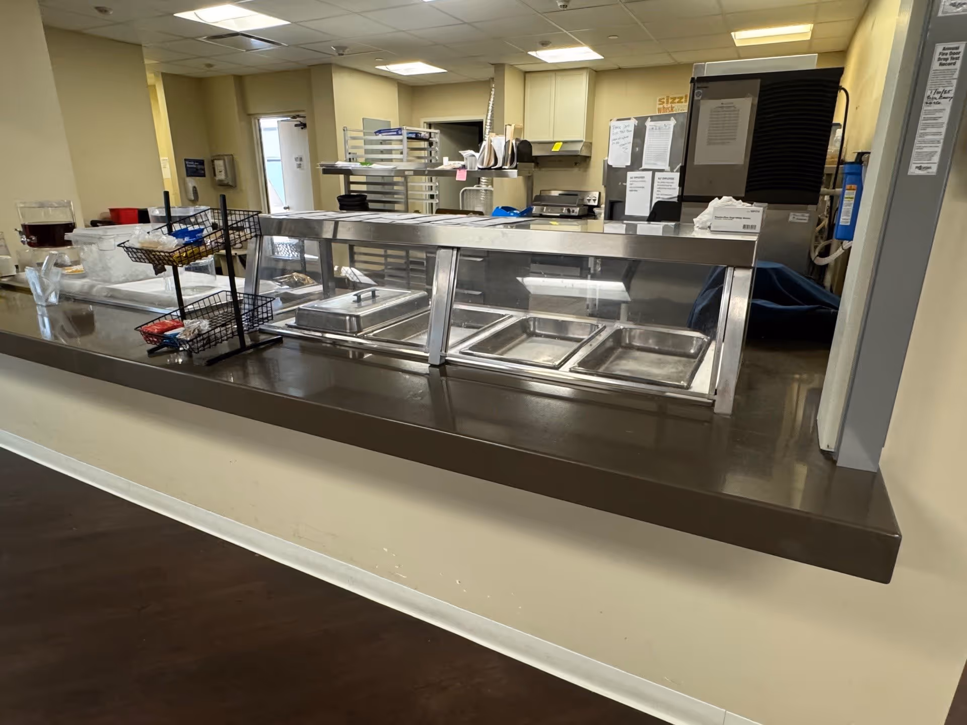 Empty cafeteria serving line with stainless steel food pans behind a glass sneeze guard in a senior living facility kitchen.
