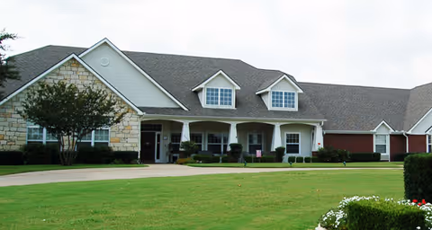 Exterior view of a single-story senior living facility building with a combination of stone and siding facade, multiple windows, and a well-maintained lawn with shrubs and flowers in front.
