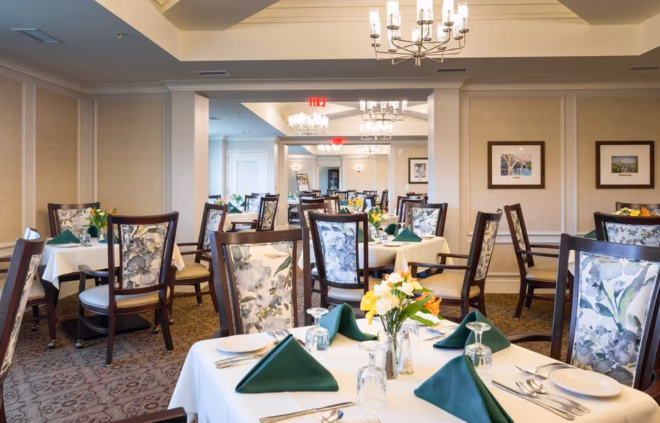 Dining room with multiple tables set with white linens, green napkins, floral centerpieces and patterned chairs.