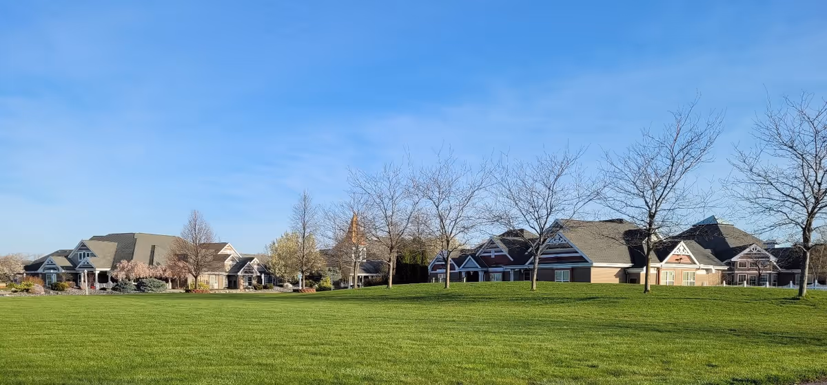 Wide view of a senior living facility named Woodcare with multiple connected buildings featuring pitched roofs and decorative trim. The buildings are surrounded by a large, well-maintained green lawn and several leafless trees under a clear blue sky.