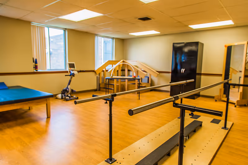 A rehabilitation room with wooden flooring featuring parallel bars for walking exercises, a padded therapy table, an exercise bike, a black storage cabinet, a large mirror, and wooden therapy equipment near two windows.