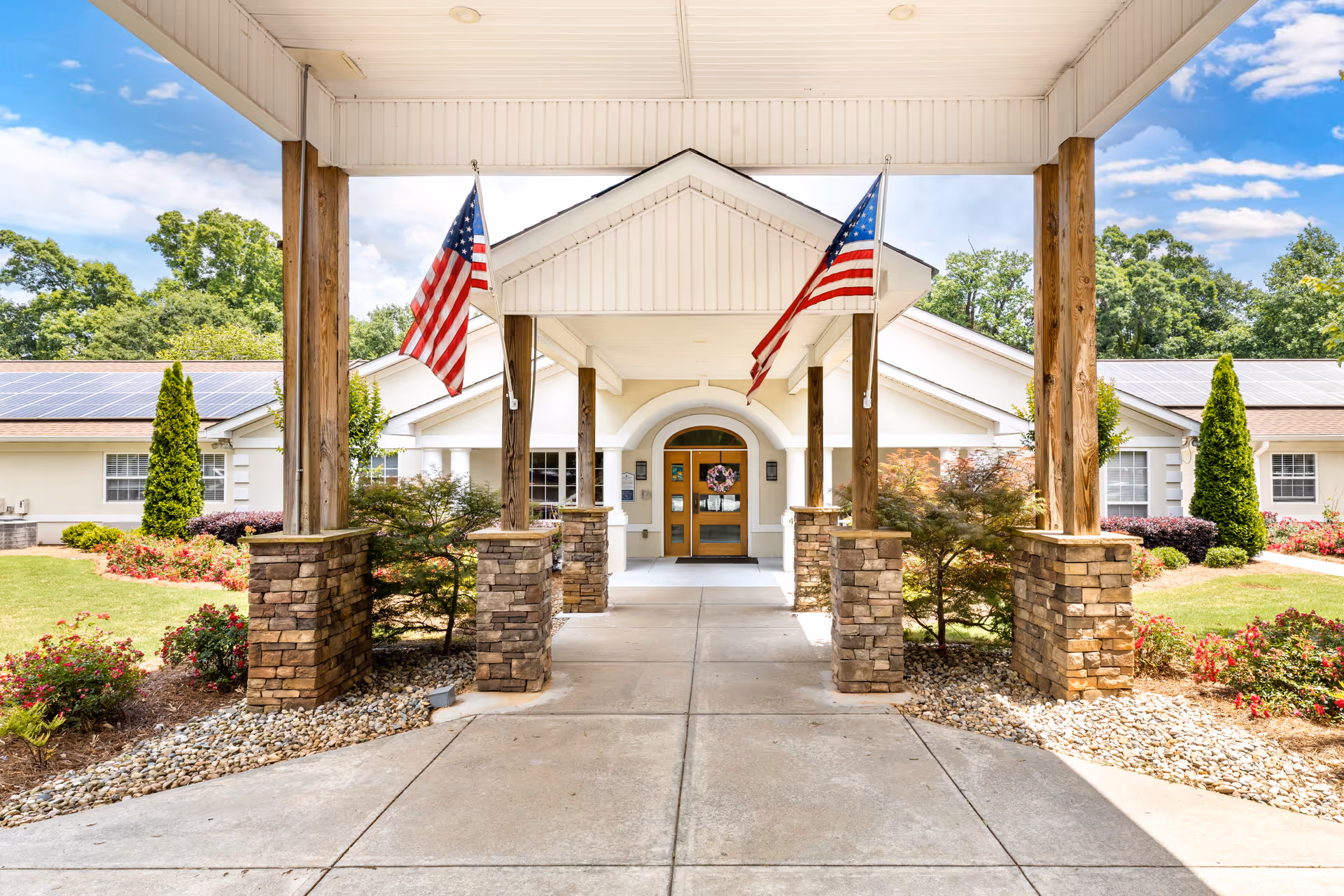 Entrance of a building with a covered walkway supported by wooden pillars with stone bases, two American flags hanging from the ceiling, landscaped garden areas with bushes and flowers on either side, and a clear blue sky with some clouds in the background.