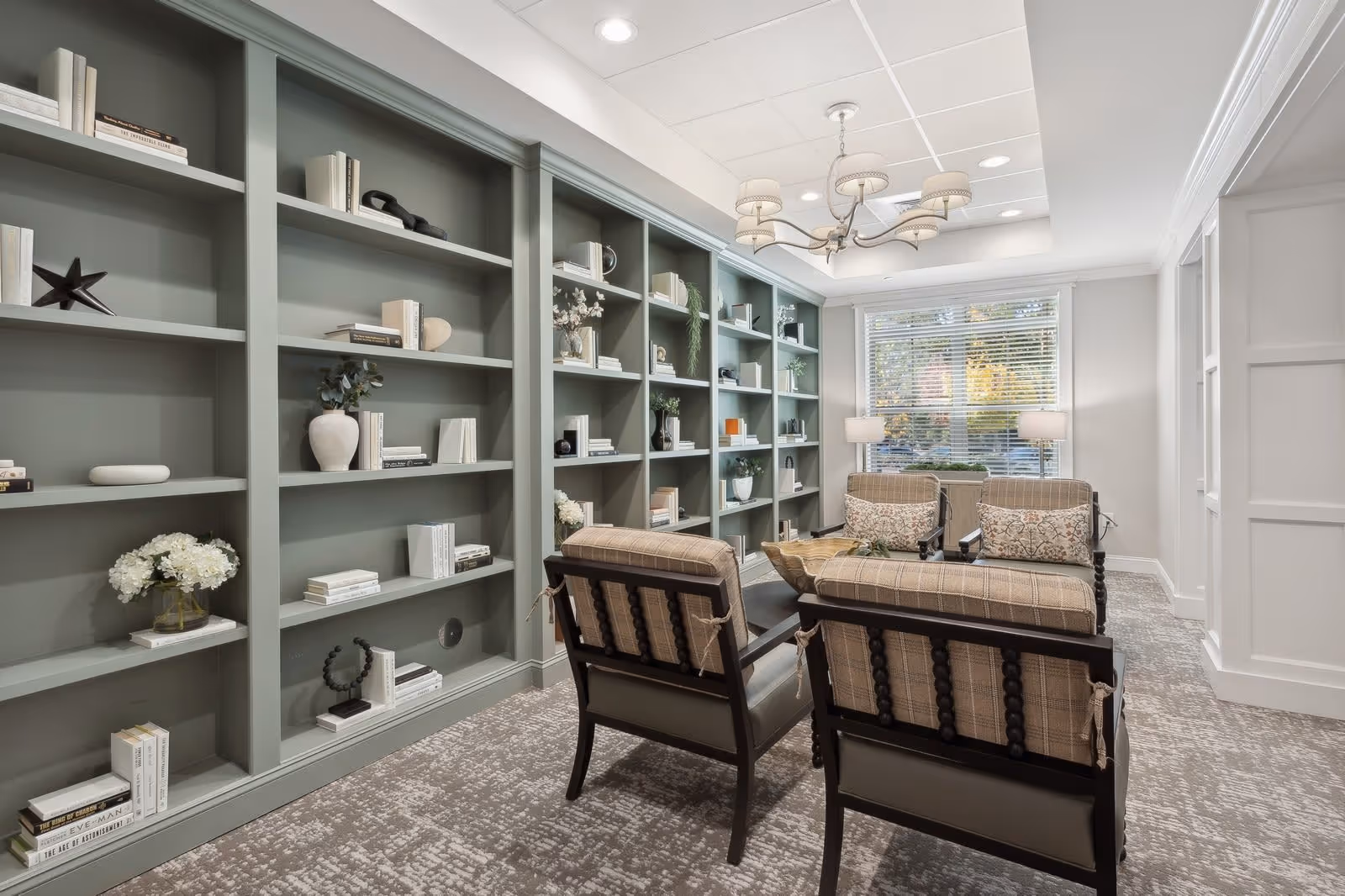 A cozy sitting area in a senior living facility with four cushioned chairs arranged around a wooden coffee table. The room features built-in green bookshelves filled with decorative items and books, a large window with blinds letting in natural light, two floor lamps, and a chandelier hanging from the ceiling.