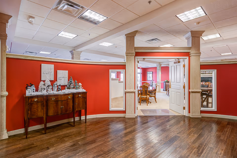 Bright interior common area with red walls, a wooden sideboard decorated for the holidays, and open doors leading to a dining room.