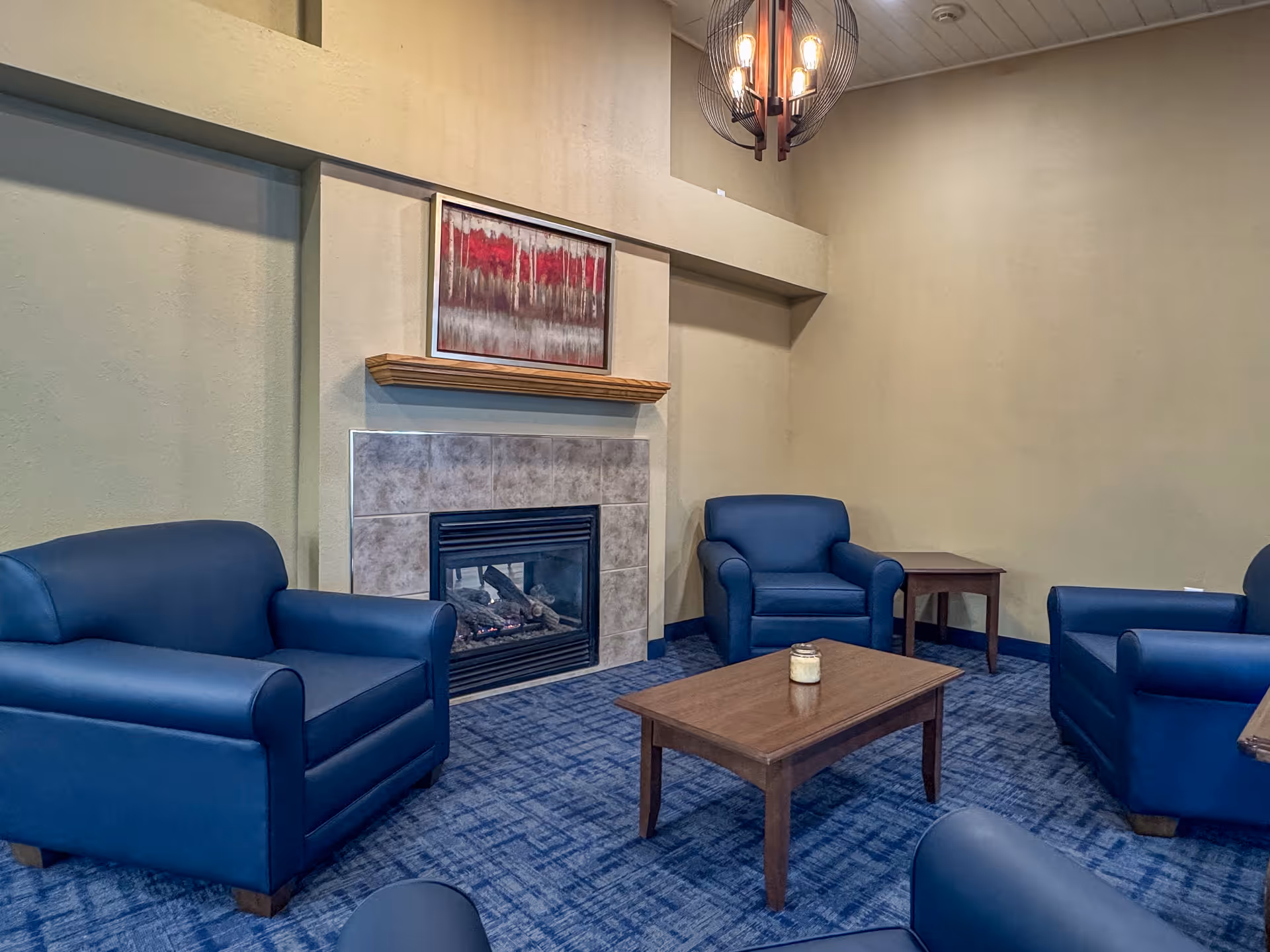A cozy living room area with four blue armchairs arranged around a wooden coffee table with a candle on it. There is a fireplace with a tiled surround and a wooden mantel above it, featuring a framed artwork with red and gray tones. The room has beige walls and blue patterned carpet, with a modern hanging light fixture above.