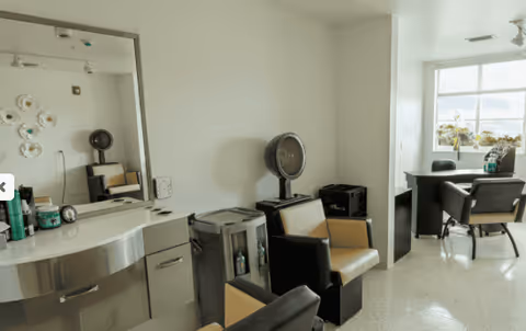 Interior view of a salon area in a senior living facility with salon chairs, a hair dryer, a large mirror, and a window letting in natural light. There are hair care products on the counter and a small table with chairs near the window.
