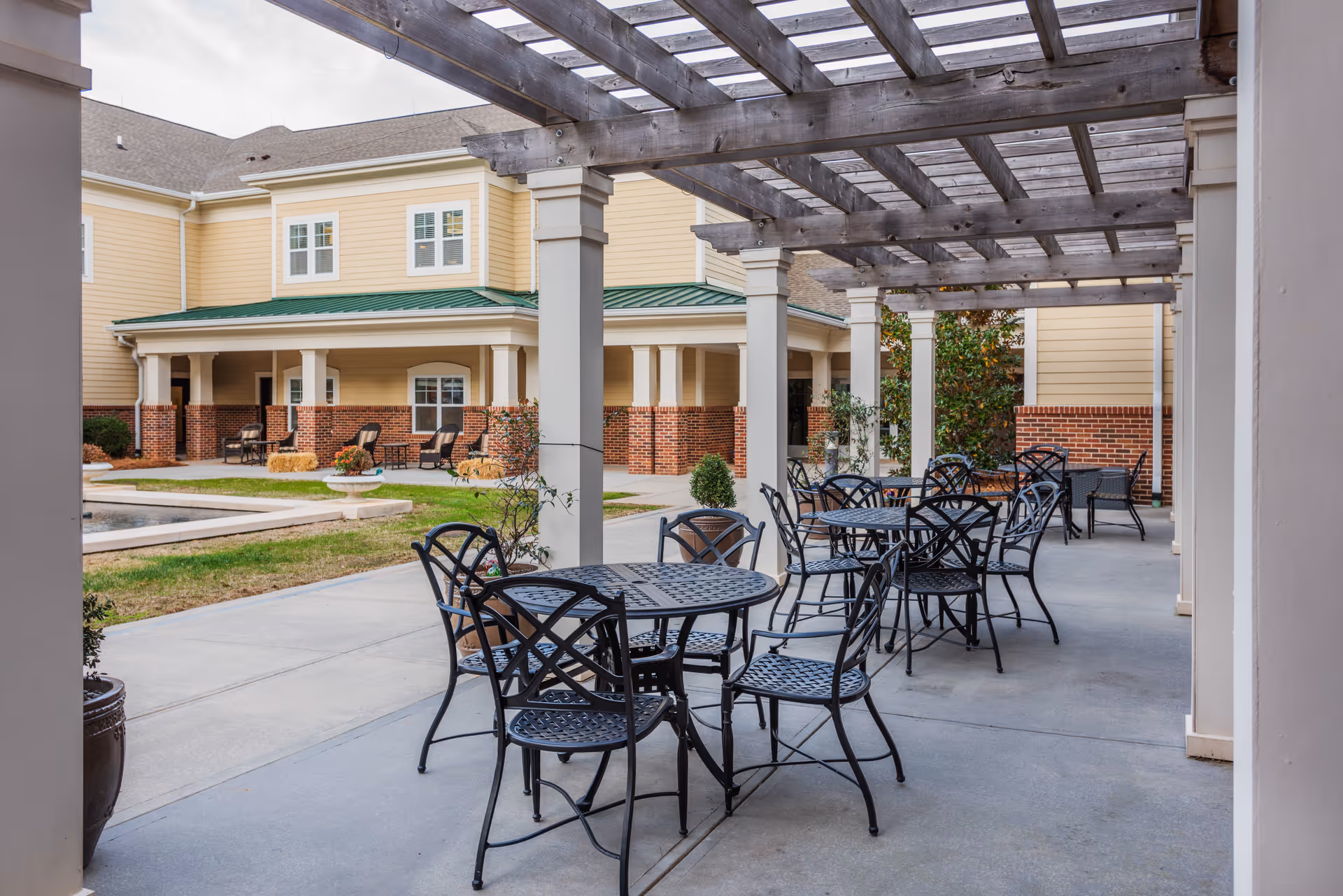 Outdoor patio area with black metal tables and chairs under a wooden pergola, adjacent to a yellow building with a green roof and brick accents. The patio overlooks a small grassy area and a water feature.