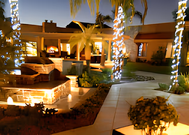 Nighttime outdoor view of a senior living facility courtyard with illuminated palm trees wrapped in white lights, a stone water feature with small fountains, a curved walkway, and a building with large windows and warm interior lighting in the background.