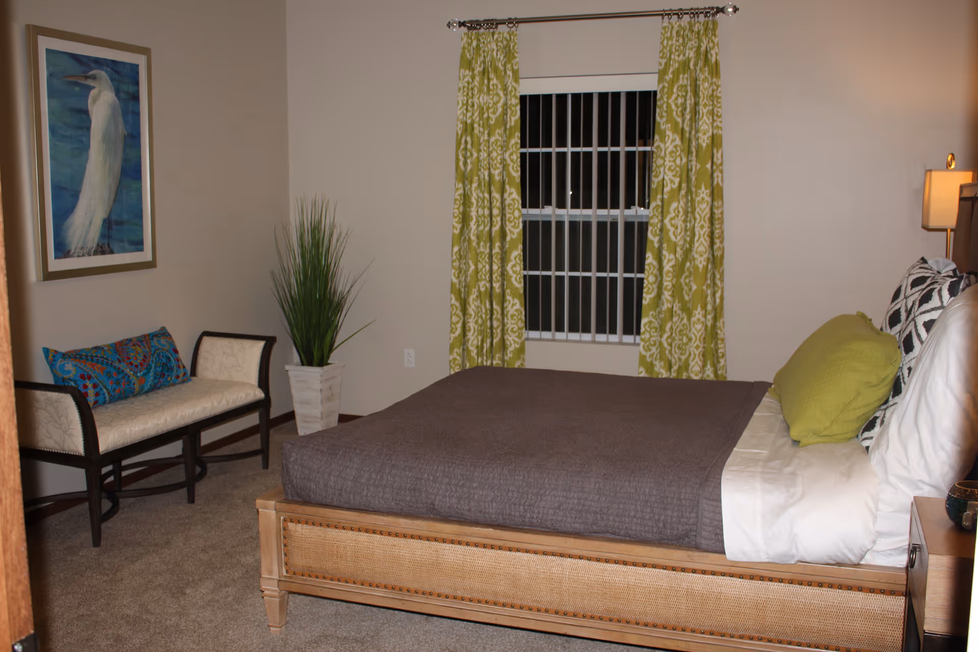 A bedroom with a wooden bed frame, gray bedspread, white sheets, and green and patterned pillows. There is a window with green patterned curtains behind the bed. To the left, there is a cushioned bench with two colorful pillows and a tall potted plant. A framed picture of a white bird hangs on the wall above the bench.