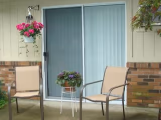 Outdoor patio area with two beige chairs and a small white metal table holding a potted plant with purple flowers. A hanging pot with pink flowers is mounted on the wall next to a sliding glass door with vertical blinds. The wall is partially brick and partially beige siding.