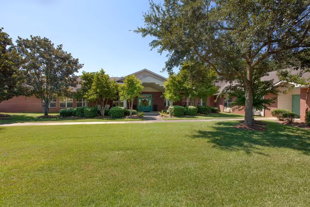 Front exterior view of Knollwood Pointe Assisted Living and Memory Care building with a well-maintained lawn, several trees, and a clear blue sky.