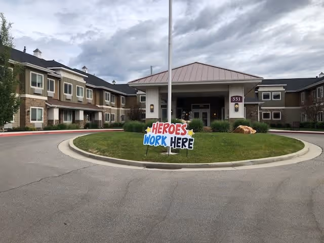 Front exterior view of a senior living facility building with a circular driveway and a grassy island in the center. A sign on the grass reads 'HEROES WORK HERE'. The building has two stories with multiple windows and a covered entrance.