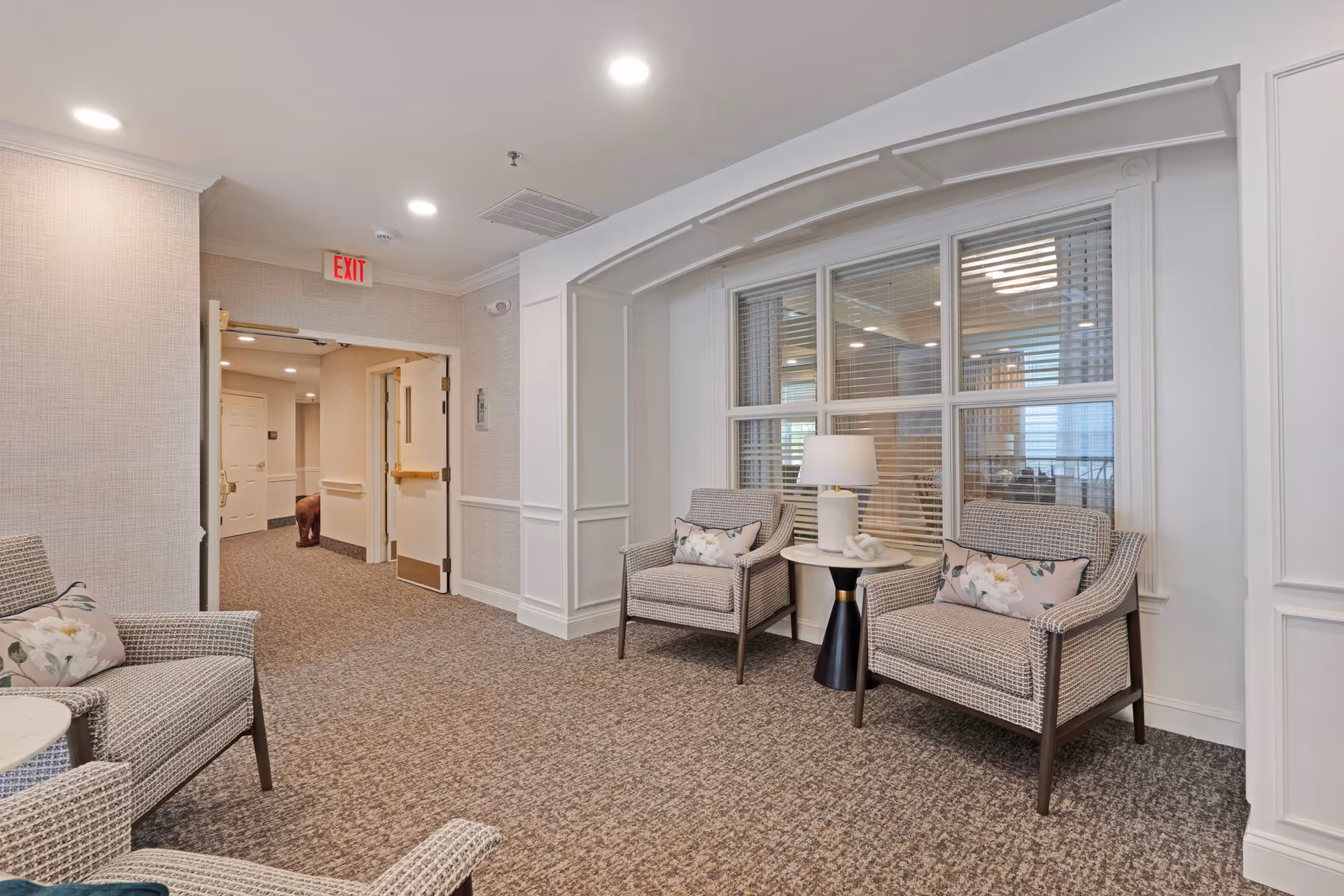 A cozy seating area in a senior living facility with two cushioned armchairs featuring floral pillows, a small round table with a lamp, and a window with blinds behind the chairs. The area has carpeted flooring, light-colored walls with decorative molding, and an open doorway leading to a hallway with an exit sign above.