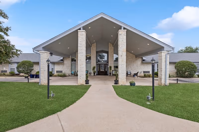 Front exterior view of a single-story building with a large covered entrance supported by stone pillars. The building is surrounded by green grass and has a paved walkway leading to the entrance. There are two black lamp posts on either side of the walkway and some bushes near the building.