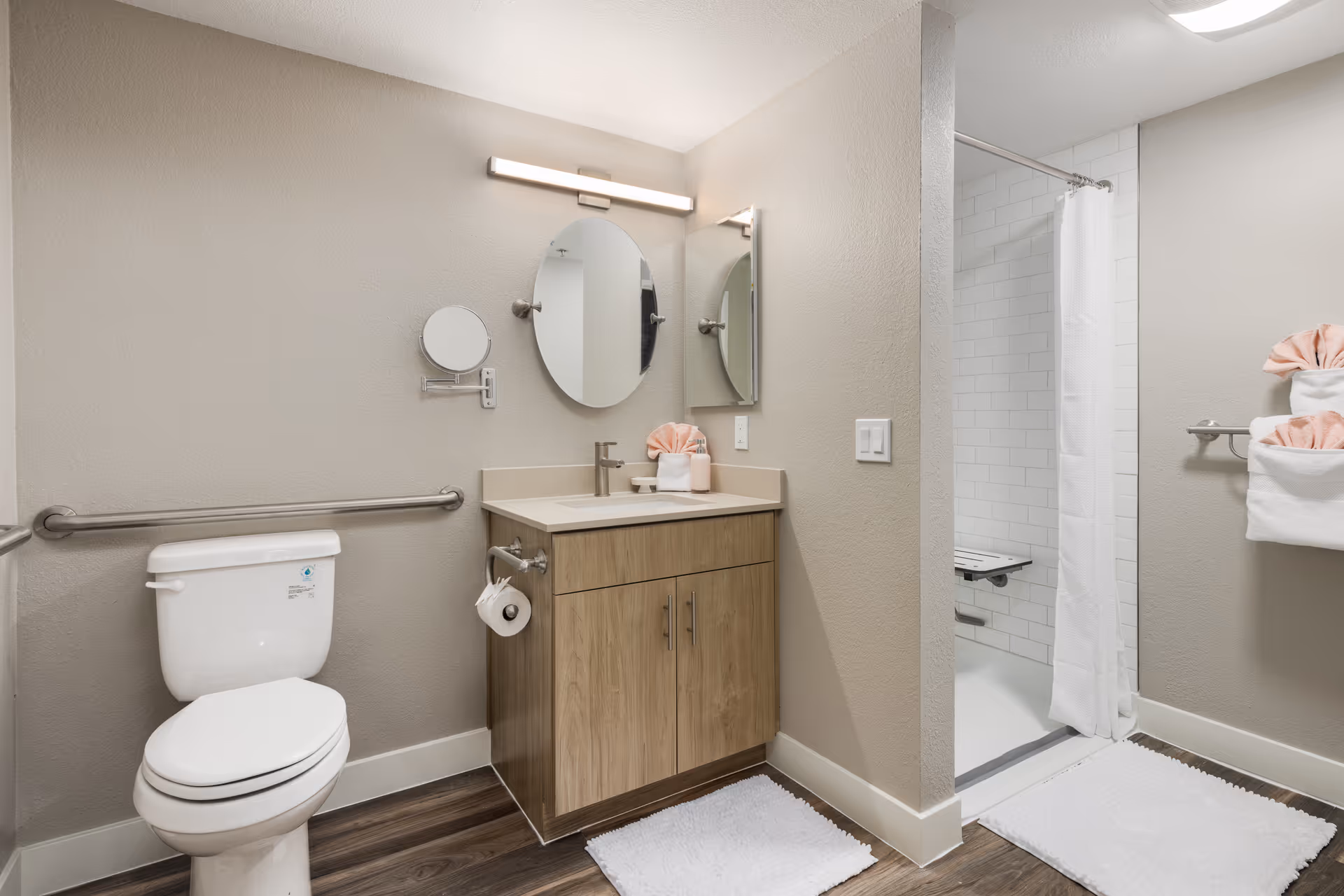 Accessible bathroom with a toilet, wooden vanity and sink, round mirror, and a walk-in shower with a curtain.